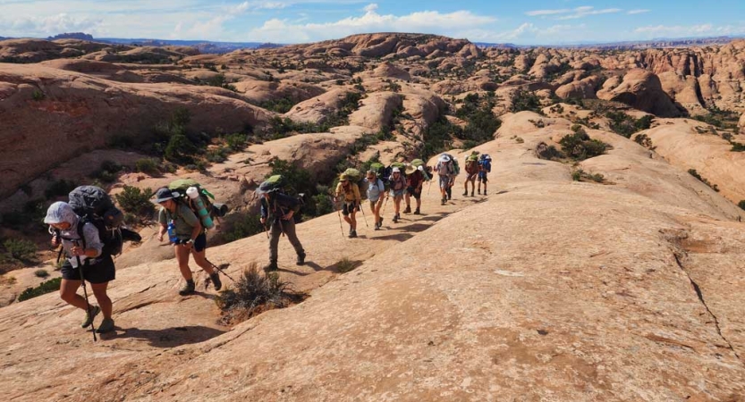 A group of people carrying backpacks hike in a line across a vast desert landscape. 